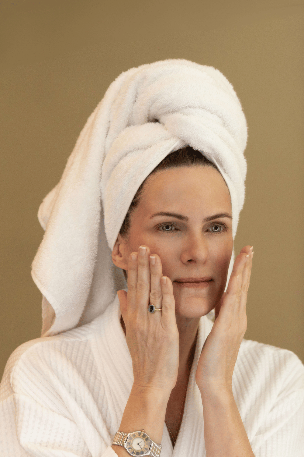 Woman with towel around hair looking at her skin in the mirror