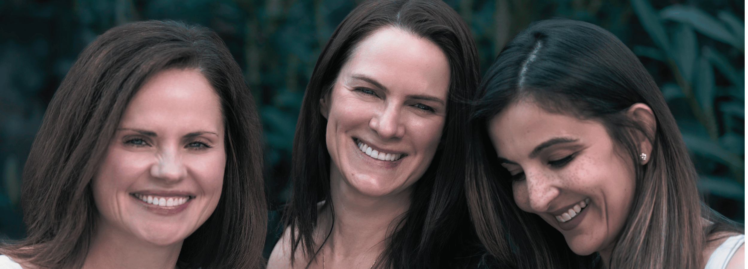 Three smiling women with long brown hair standing closely together outdoors against a dark green blurred background.