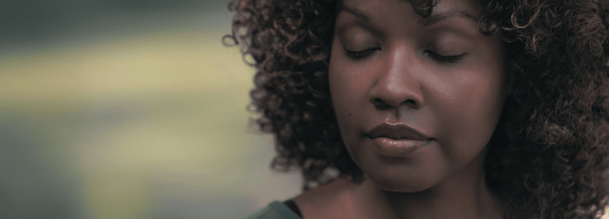 A close-up portrait of a woman with curly dark hair, her eyes closed, set against a soft, blurred green and gray background.