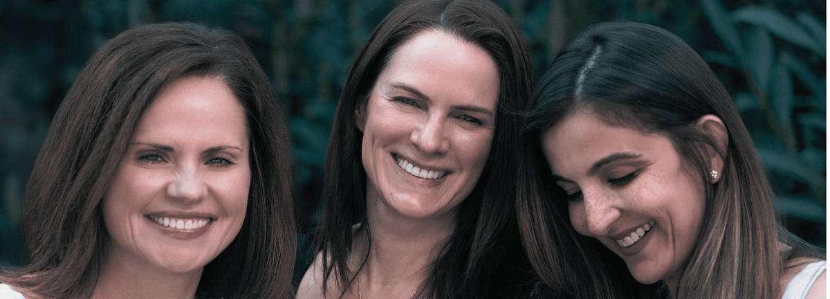 Three smiling women with long brown hair standing closely together outdoors against a dark green blurred background.